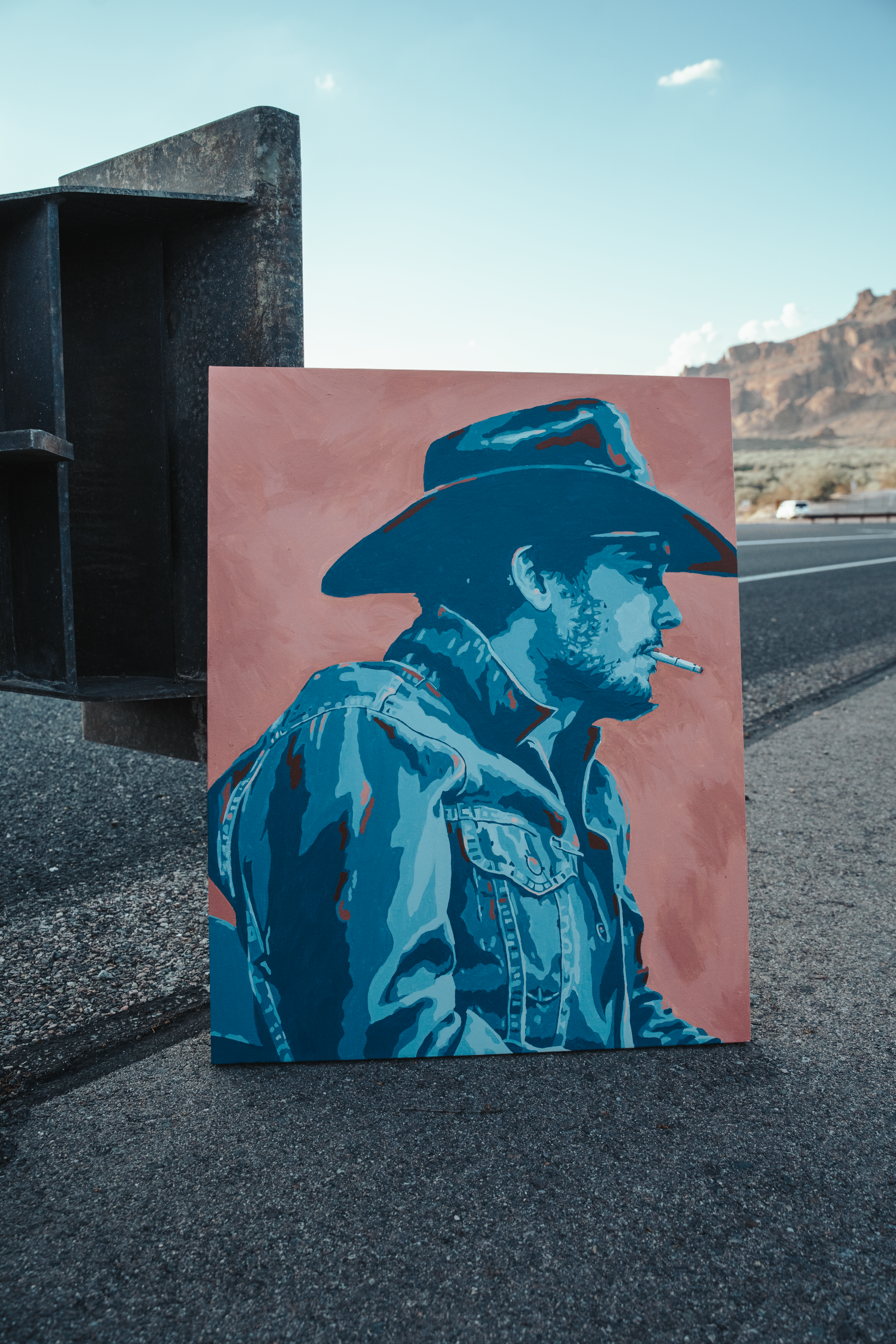 A finished painting leaning against a black steel structure beside a desert highway, mountains in the distance